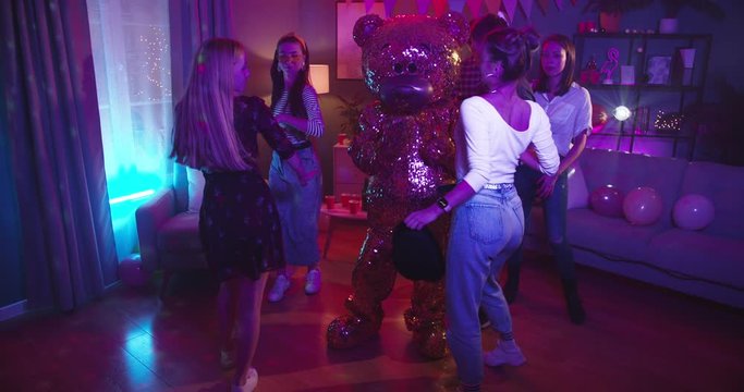 Home Party In The Decorated Cozy Living Rom With Blue And Red Lights - Yung Girls And Guys Dancing And Having Good Time With Big Golden Life-size Doll Of Teddy Bear. Indoors.