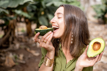 Young smiling woman eating avocado outdoors, standing on the tropical plantation. Concept of...