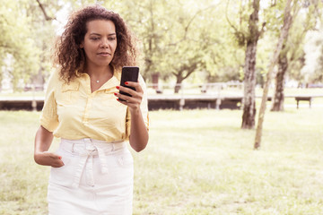 Serious woman reading news on smartphone in city park. Young lady wearing casual shirt and skirt and standing with blurred green trees and lawn in background. Communication concept. Front view.