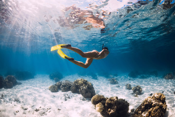Woman glides underwater with yellow fins over sandy sea and coral.