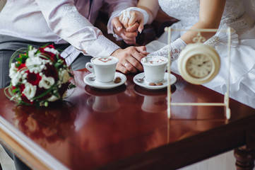 The bride and groom tenderly hold hands, sitting in a cafe with cups of coffee with foam in the form of a heart, and a bouquet of roses and eustomas. Photography, concept.
