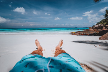 POV of male wearing swimming shorts with tanned legs on paradise white sand tropical exotic beach with view to turquoise blue ocean. Travel holidays vacation concept - obrazy, fototapety, plakaty