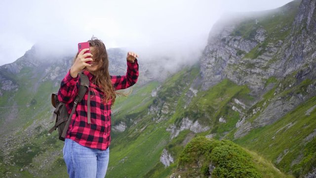 Pretty girl backpacker taking selfie in mountain. Woman in a red checkered shirt on top, smiling and posing, looking at camera while standing against picturesque view peaks covered clouds