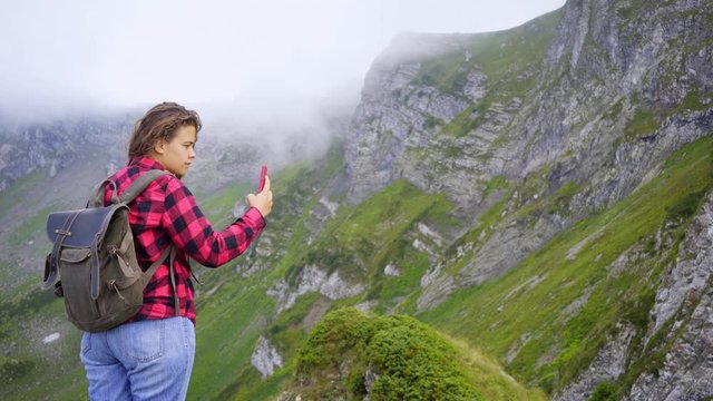 Pretty girl backpacker taking selfie in mountain. Woman in a red checkered shirt standing on top, taking selfie or sharing video via smartphone while enjoying picturesque view peaks covered clouds