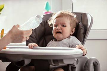 Beautiful baby in the child dining chair drink milk from the bottle in the kitchen. Crying baby before the lunch. Mother's hands. Lifestyle. Sunny day.