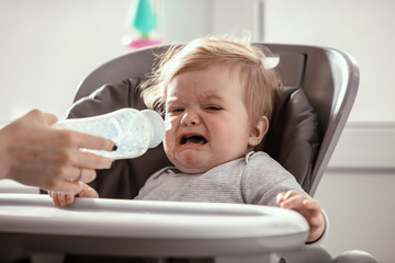 Beautiful baby in the child dining chair drink milk from the bottle in the kitchen. Crying baby before the lunch. Mother's hands. Lifestyle. Sunny day.