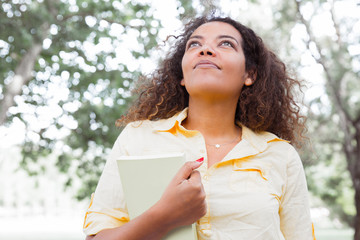 Dreamy young woman holding book in city park. Woman looking upwards and standing with blurred trees in background. Summer and education concept. Front view.