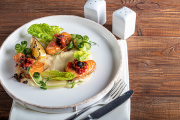 Celery and potato puree with pieces of red fish, garnished with herbs and fresh cucumbers. On a white plate with lemon slices. On a wooden table.
