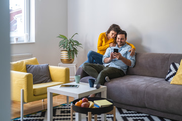 Couple using mobile phone at home stock photo. Positive couple is surfing internet on smart phone while sitting on sofa - Powered by Adobe