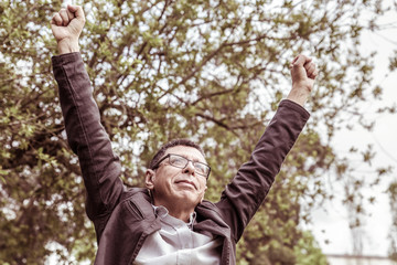 Cheerful middle-aged man raising arms in park. Guy wearing casual clothes and celebrating success with green trees in background. Achievement and nature concept. Front view.