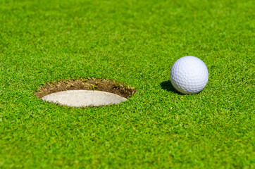 A hole over golf ball on green. Shallow depth of field. Focus on the ball and the hole.