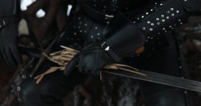 Cosplayer portrays a character of medieval warrior and cleans his sword by dry grass at winter forest.