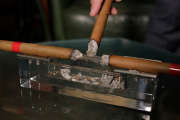 two smoking cigars lie in a transparent ashtray, a man brings a third cigar to an ashtray to knock down the ashes