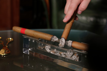 two smoking cigars lie in a transparent ashtray, a man brings a third cigar to an ashtray to knock...