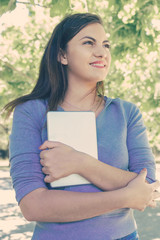 Fototapeta premium Dreamy pretty young lady holding tablet computer in park. Beautiful woman wearing hoodie, standing and looking away with green trees in background. Leisure and nature concept. Front view.