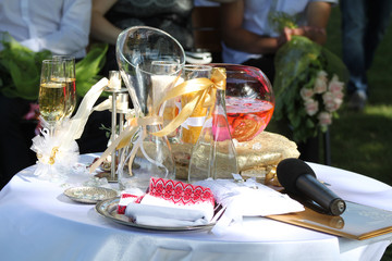 wedding rings on a small pillow, rose petals floating in pink water in a glass spherical vase, white and yellow sand in glasses, two glasses with champagne, a microphone