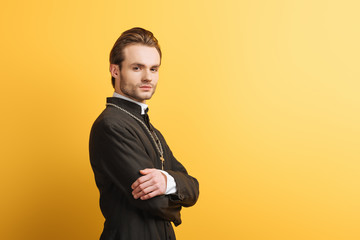confident catholic priest with crossed arms looking at camera isolated on yellow
