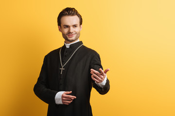 smiling catholic priest standing with outstretched hand while looking away isolated on yellow