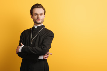 confident catholic priest standing with crossed arms and looking away isolated on yellow - obrazy, fototapety, plakaty