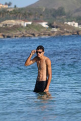 Attractive shirtless athletic young man on the beach by the sea. Young man in shorts and sunglasses walking by the sea coast. Travel fashion. Sport lifestyle