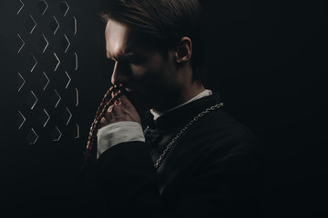 young thoughtful catholic priest kissing wooden rosary beads near confessional grille in dark with...