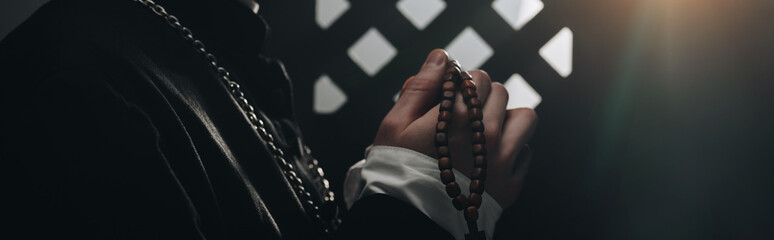 Partial view of catholic priest holding wooden rosary beads near confessional grille in dark with rays of light, panoramic shot