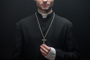 Fototapeta na wymiar cropped view of catholic priest touching silver cross isolated on black