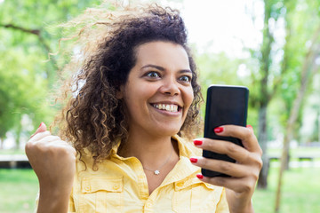 Cheerful woman reading news on smartphone in city park. Young lady pumping fist and standing with blurred green trees and lawn in background. Success concept. Front view.