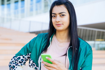 Medium shot of pretty young mixed-race woman in green jacket and rose T-shirt standing on stairs, looking confident, looking at camera, holding phone in hand. Lifestyle concept