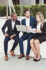 Hardworking business group checking reports outside. Three managers sitting near office building, using laptop, holding documents and talking. Corporate meeting outside concept