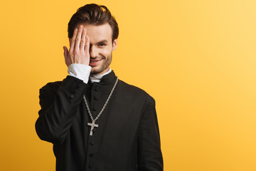 smiling catholic priest covering eye with hand while looking at camera isolated on yellow