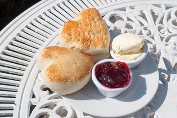 Pair of fresh heart-shaped English scones sitting together outdoors on a sunny decorative table with a dollop of Cornish clotted cream and strawberry preserves