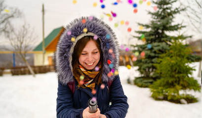 Fototapeta na wymiar Emotional woman pops confetti petard and opens her mouth with delight. Adult female in hooded jacket admires and pops confetti petard into camera while standing near conifer tree on winter day in