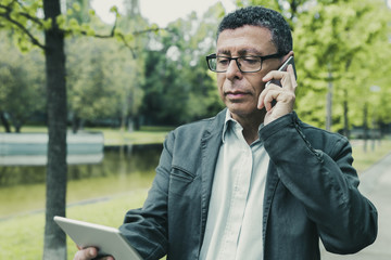 Serious man browsing on tablet and talking on phone in park. Guy wearing casual clothes and walking on pavement with green trees in background. Communication and nature concept. Front view.