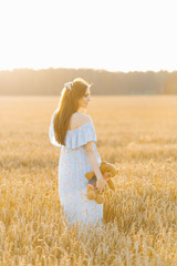 A pregnant young woman in a blue sundress stands in a field of wheat at sunset in summer and holds a stuffed bear toy in her hand