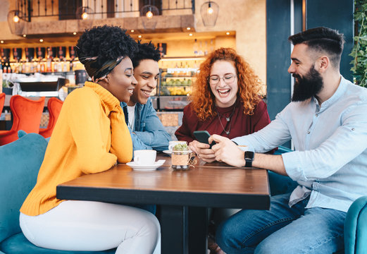 Four friends, two males and two females, sitting at a cafe, drinking coffee and talking. The bearded man is showing something on his cellphone.