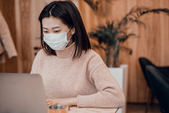 Asian woman in a protective mask sits in a cafe at a laptop. Protecting the population from coronaviruses by protecting the respiratory tract. A girl works in a coffee shop in a blue medical mask
