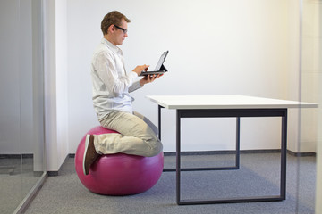Man on stability ball working with tablet at desk in the office - changing position