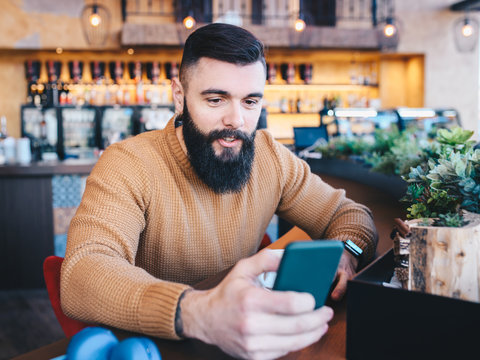 A young man with a beard looking at his cell phone in a cafe.