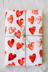 a gift wrapped in homemade wrapping paper with red hearts tied with jute thread for Valentine's day on a white table