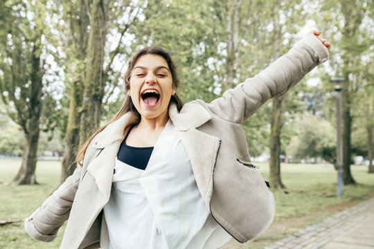 Cheerful pretty young woman spreading arms and screaming in park. Excited lady wearing jacket, looking at camera and having fun with green trees in background. Happiness concept. Front view.