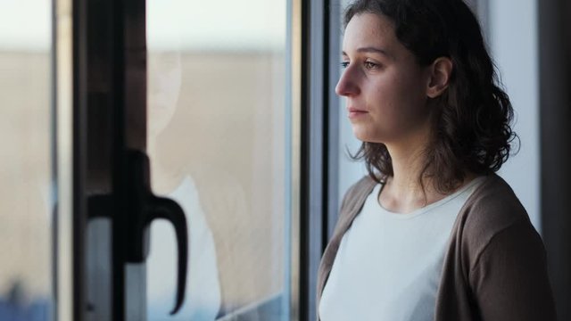 Video of sad young woman crying while looking through the window at home.
