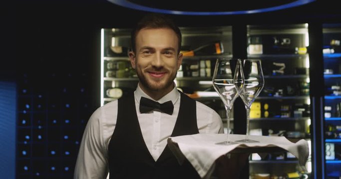 Portrait of Caucasian handsome happy male waiter smiling cheerfully to camera with tray and two empty glasses for wine. Close up of attractive man working in restaurant. Worker serving drinks.