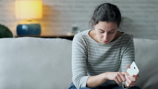 Video of worried young woman using her mobile phone while sitting on sofa in the living room at home.