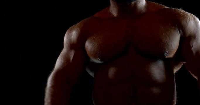 a large bald man with a beard and bronze skin poses on a black background. the bodybuilder lifts up his arms, bends them, and strains his muscles