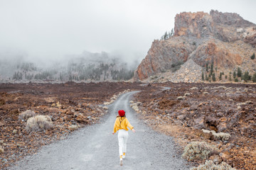 Picturesque rocky mountains in the clouds with dirt road on the volcanic valley. Woman in bright clothes walking on the road, wide view