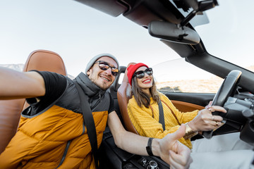 Selfie portrait of a happy couple while driving convertible sports car on the desert road during a...