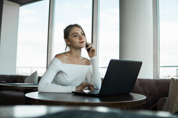 Woman working on laptop at office while talking on phone, backlit warm light. Portrait of young smiling business woman calling her best friend, having break, telling something funny, sitting in cafe.