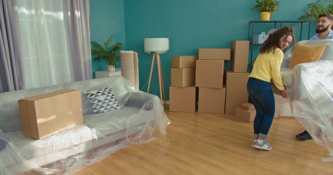 Caucasian good looking joyful man and woman carrying big armchair in living room when moving in. Happy just-married couple bringing packed furniture to apartment with boxes when removing.