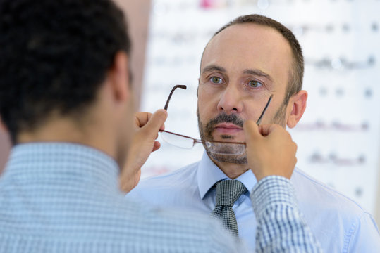 Young Male Optician Fitting Glasses Onto Mature Male Customer